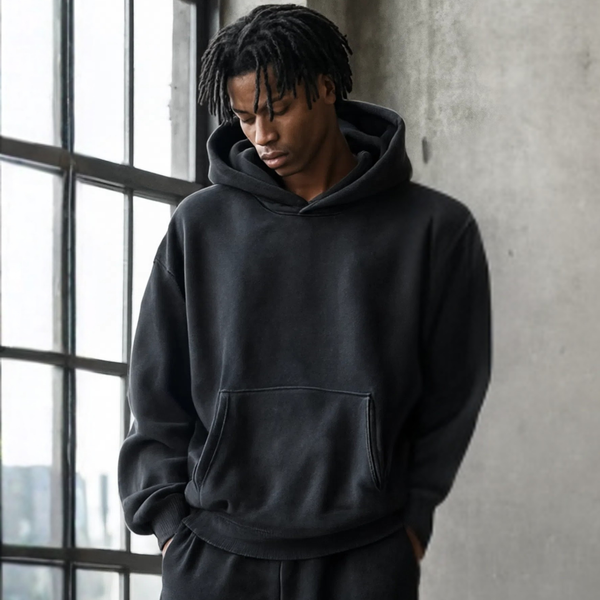 Person wearing a black hoodie standing against a concrete wall with large windows.